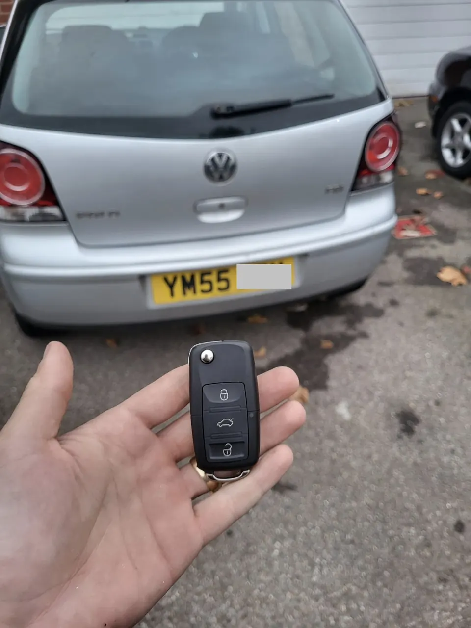 Three-button Volkswagen flip remote key held in front of a silver Volkswagen Polo