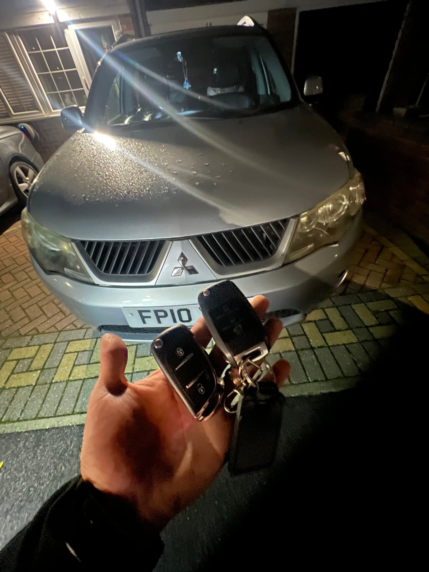 Two remote keys held in front of a silver Mitsubishi Outlander on a driveway at night