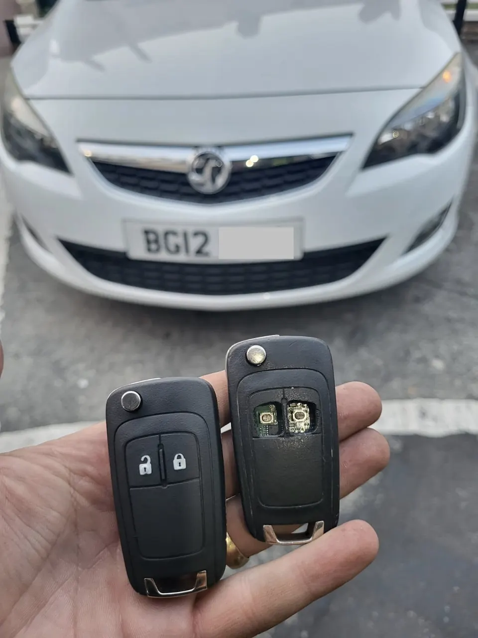 Two Vauxhall flip remote keys, one opened to show the circuit board, in front of a white Vauxhall Astra