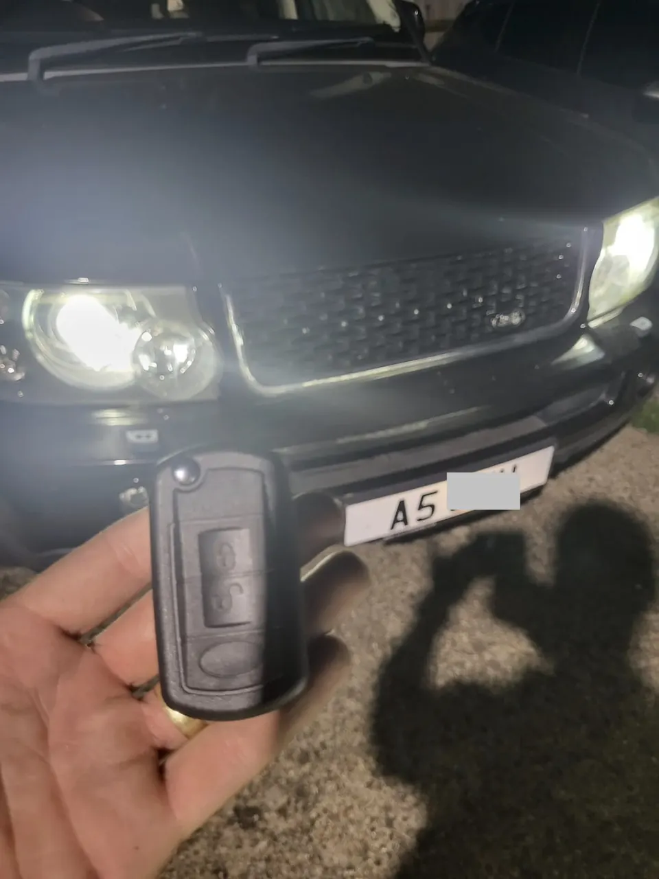 Land Rover flip remote key held in front of a Range Rover Sport with headlights on at night