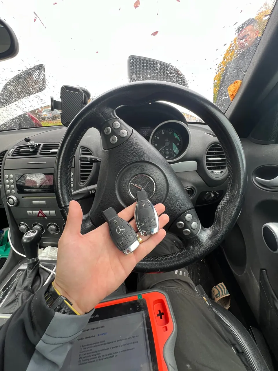 Two Mercedes keys held at the wheel of a Mercedes SLK with a diagnostic tablet on the technician's lap