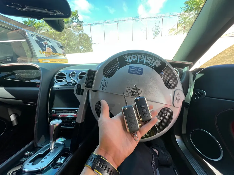 Two Bentley keys held inside a Bentley cockpit with a steering wheel lock fitted